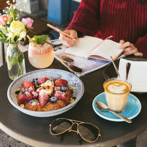 En åben notesbog, kaffe og morgenmad på et cafébord